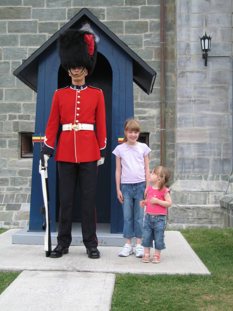 Young girl and toddler girl standing beside guard in black pants, red jacket and black fur hat at la citadelle in quebec city.