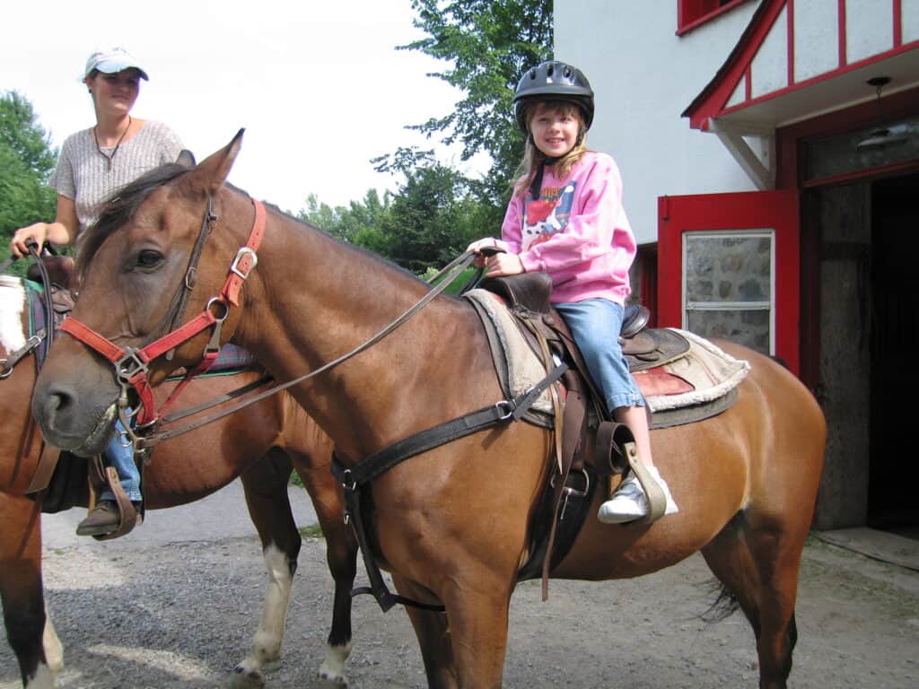 Young girl in capri jeans, pink sweatshirt wearing black helmet on brown horse and guide on horse beside her at chateau montebello in quebec, canada.