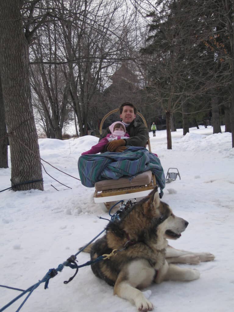 Dad and young girl bundled up on dog sled sitting in snow with one husky laying on ground in front of them.