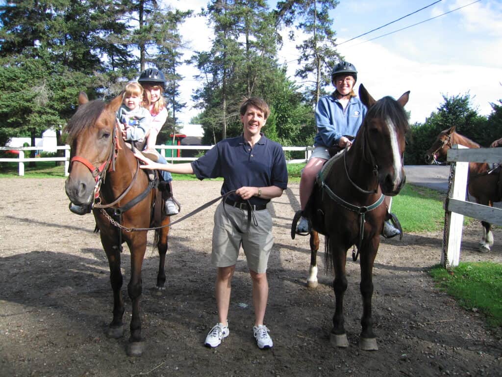 A dad standing between two brown horses holding reins with mom sitting on horse on right and young girl and toddler girl sitting on horse on left.