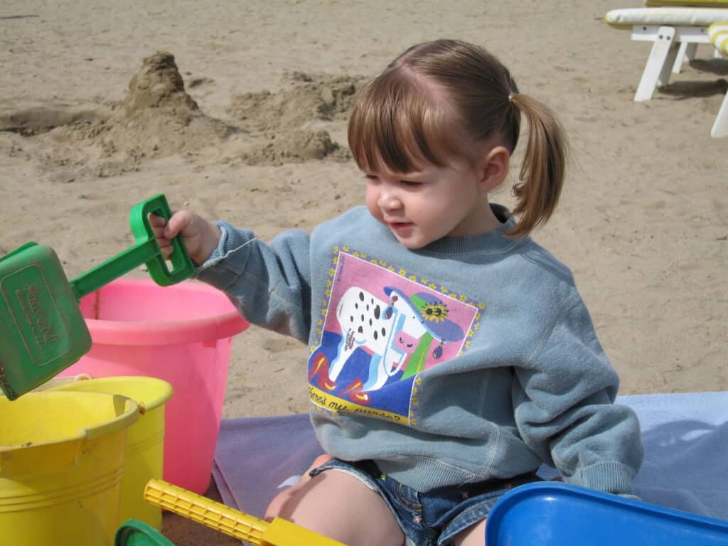 Toddler girl in sweatshirt plays in sand with colurful plastic pails and shovel at Whitefish Lake in Montebello, Quebec.