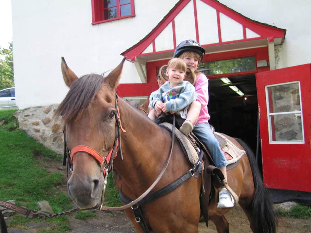 Young girl and toddler girl sitting on back of brown horse in front of red and white barn at chateau montebello in quebec.