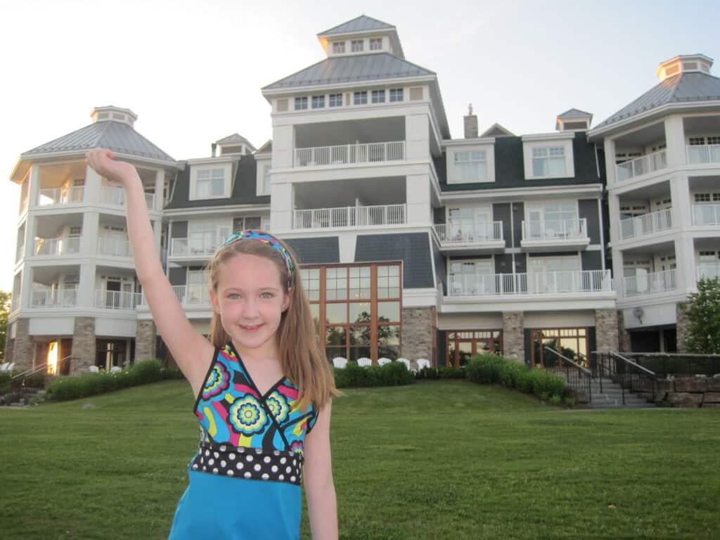 young girl wearing colourful dress with hair in matching headband poses with one arm in air standing on grass lawn in front of the Rosseau Muskoka.