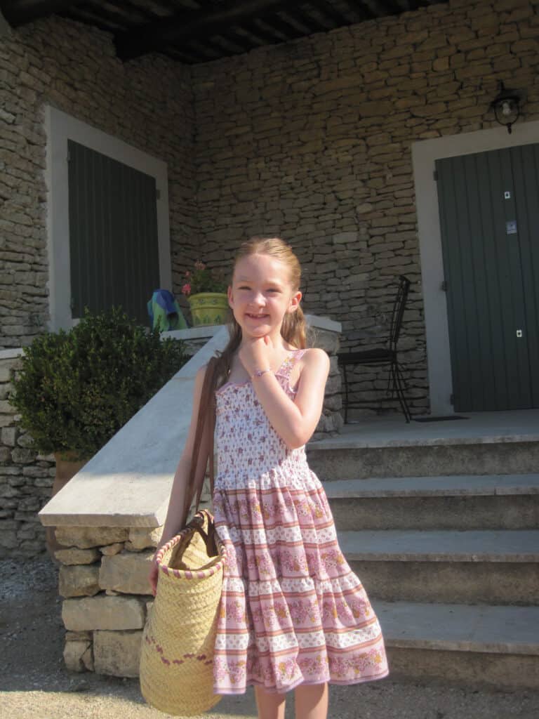 Young girl in light purple flowered dress standing on steps of stone house carrying large straw basket with leather straps.