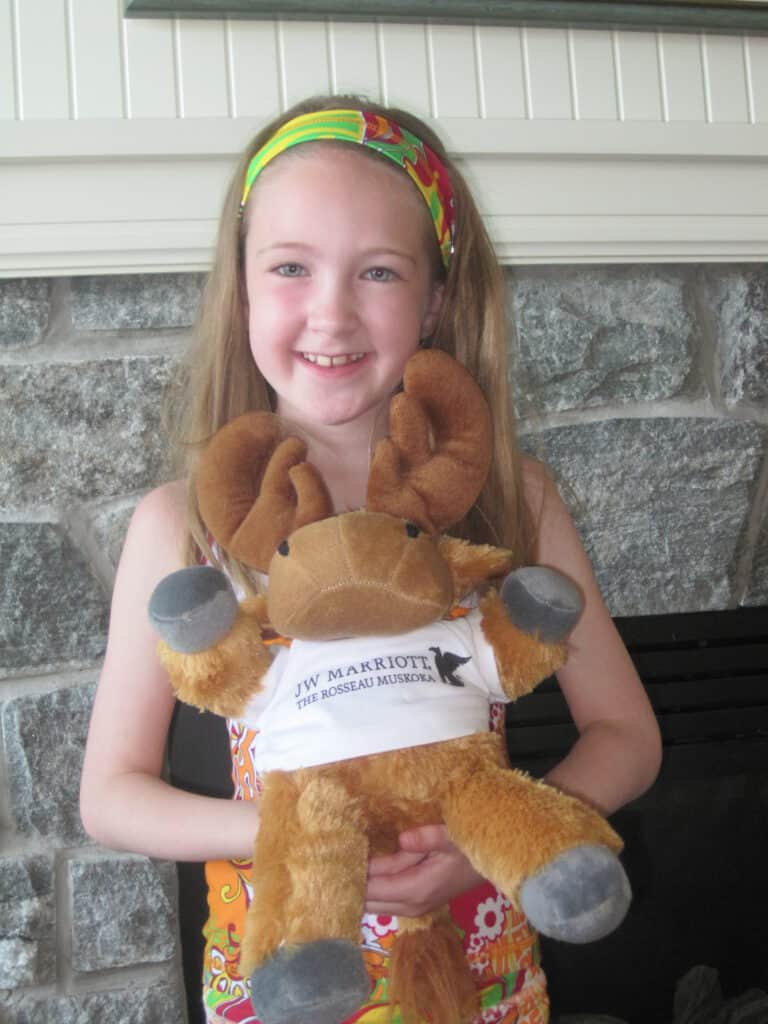 young girl standing in front of stone fireplace holding a stuffed moose wearing a JW Marriott The Rosseau Muskoka t-shirt.