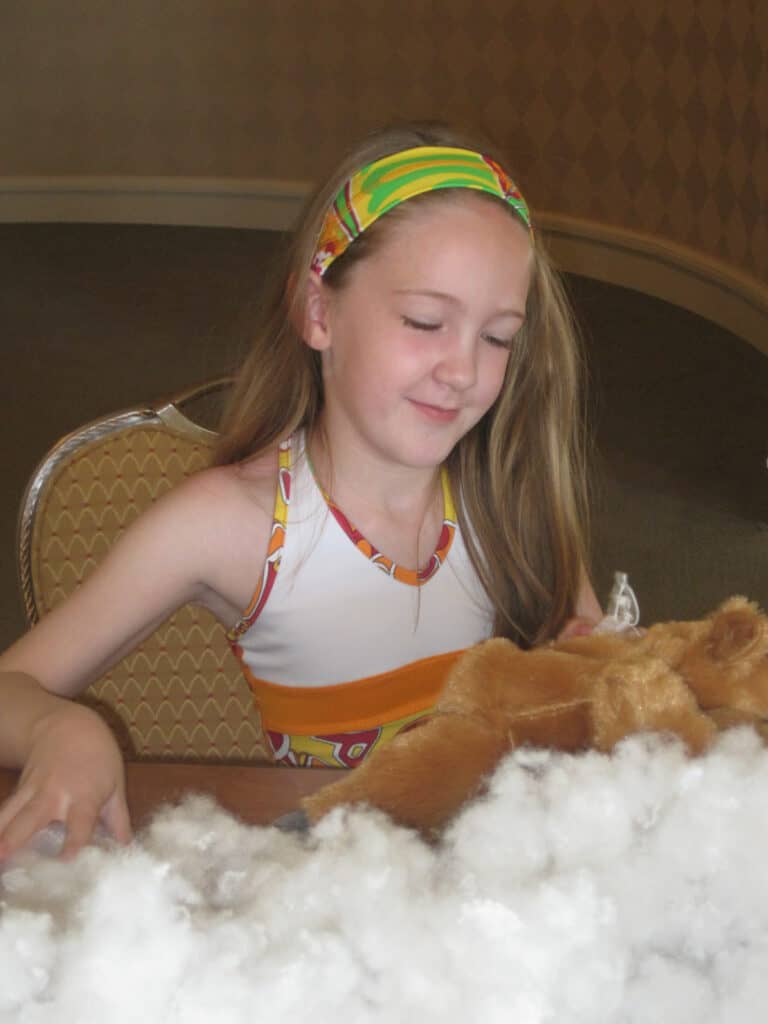 young girl in dress and matching headband sitting with stuffed animal and white stuffing on table in front of her.