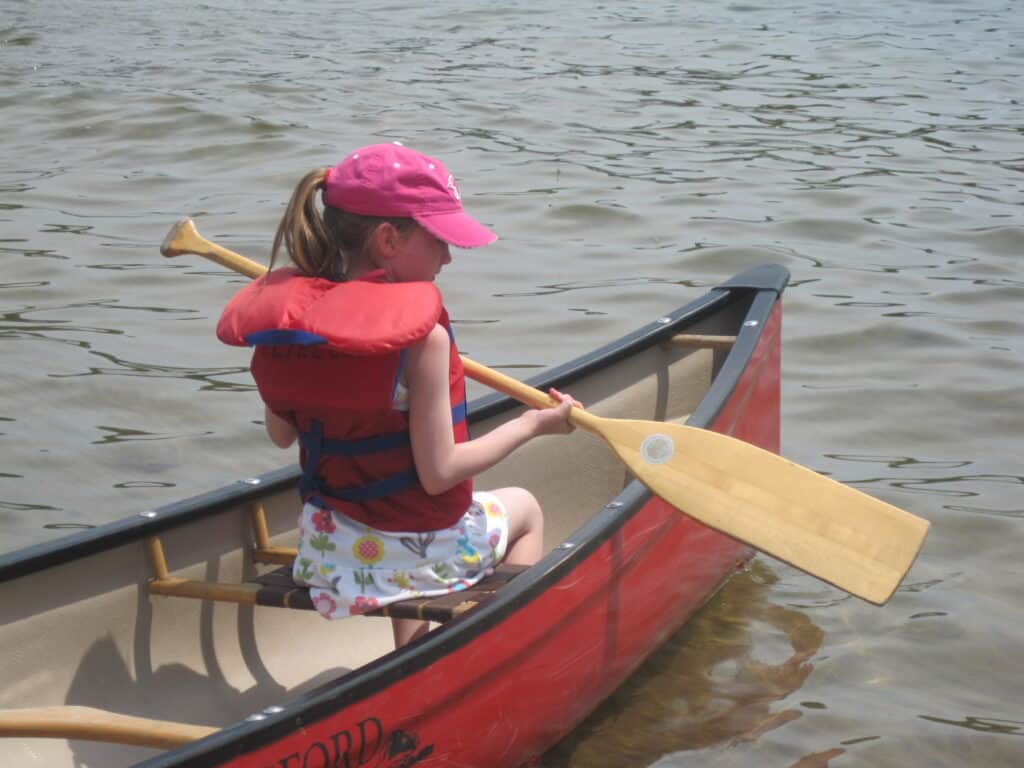 front end of red canoe in water with young girl wearing red life jacket and pink cap sitting on bench holding paddle.