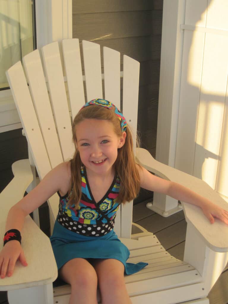 young girl in colourful dress and matching headband sitting in wooden white muskoka (adirondack) chair on the balcony of suite at the Rosseau Muskoka - Canada.