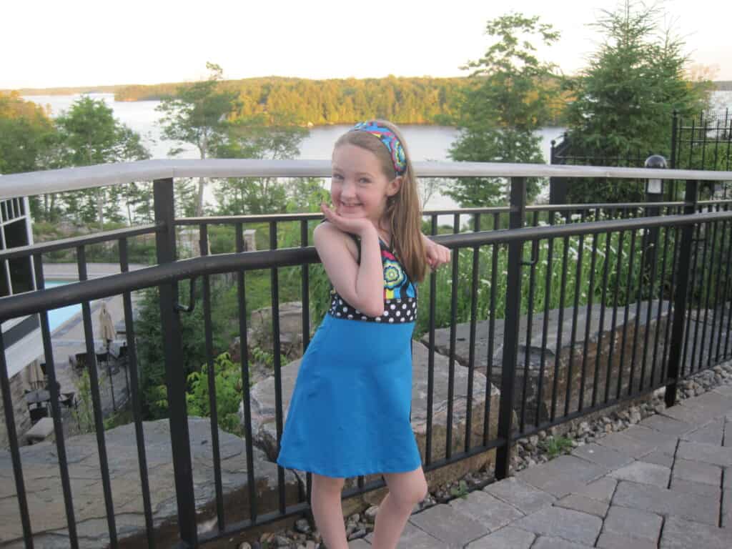 young girl in colourful dress poses with hand under chin by black railing around walkway and lake in background at rosseau muskoka.