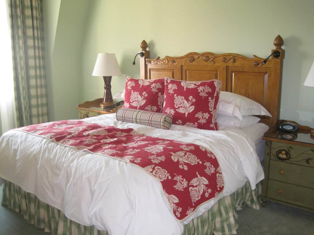 wooden bed frame and bedside tables with linens in red and green with light green walls and light green plaid curtains.