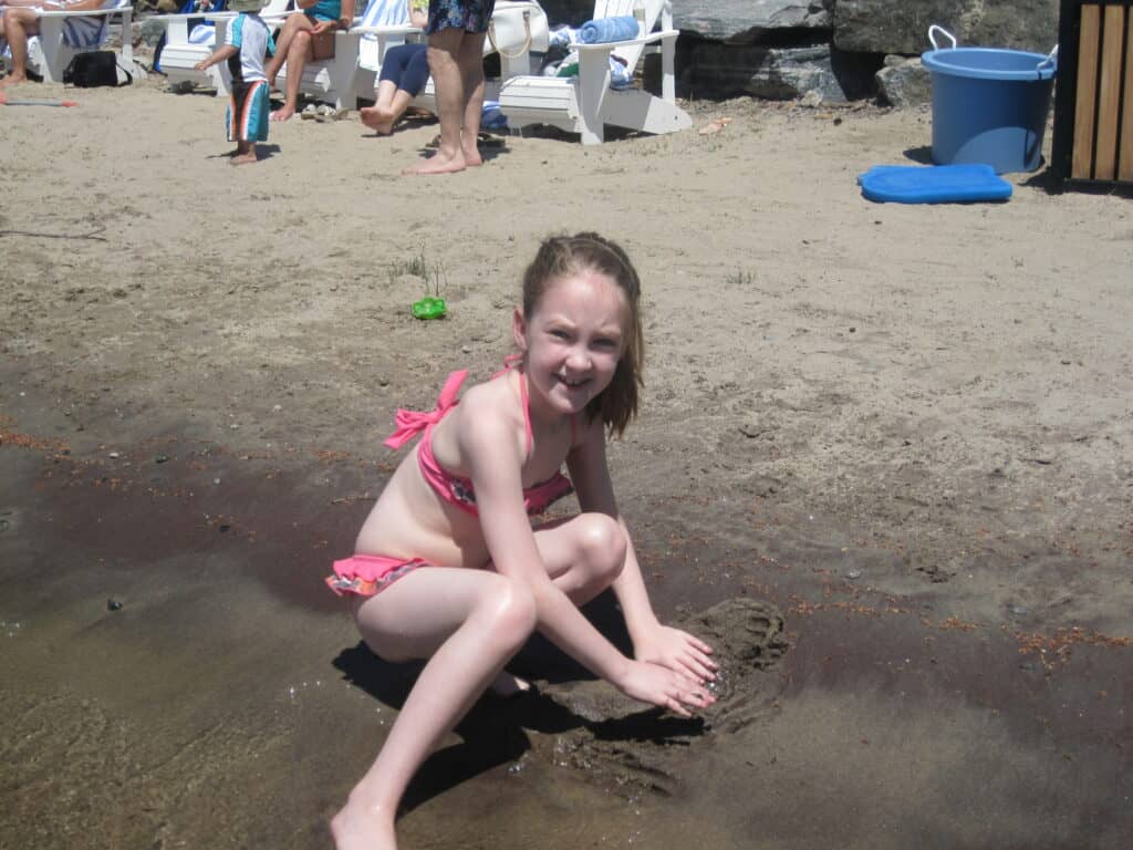 young girl in pink bikini playing on sandy beach of lake rosseau with people and white beach chairs in background - Rosseau Muskoka