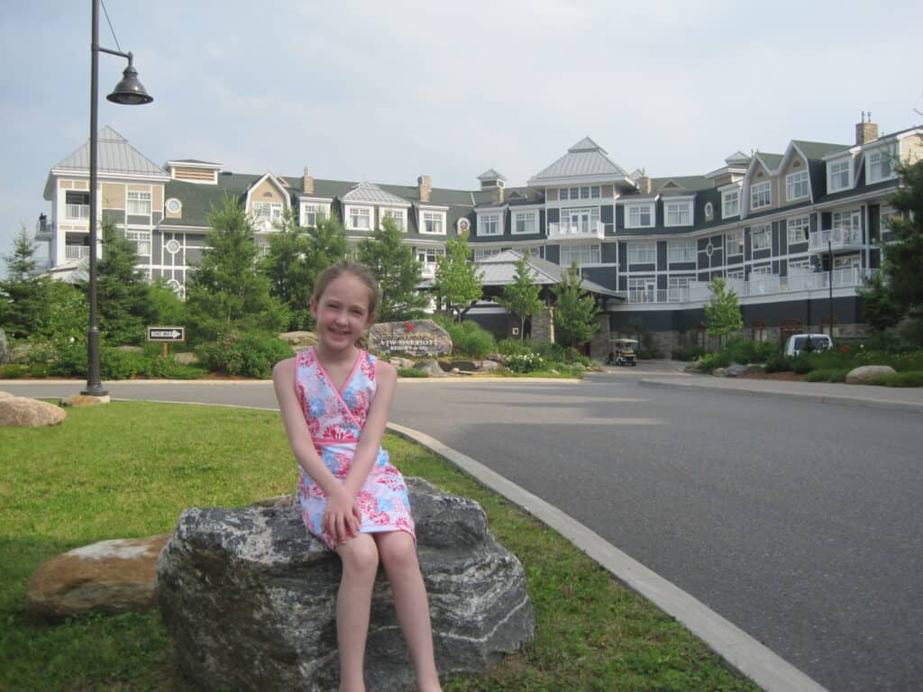 young girl with ponytail wearing pink and blue dress sitting on rock alongside driveway at entrance to the Rosseau Muskoka with main building in background.