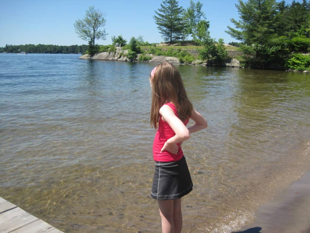 young girl in dark skirt and red tank top standing by shore of Lake Rosseau 