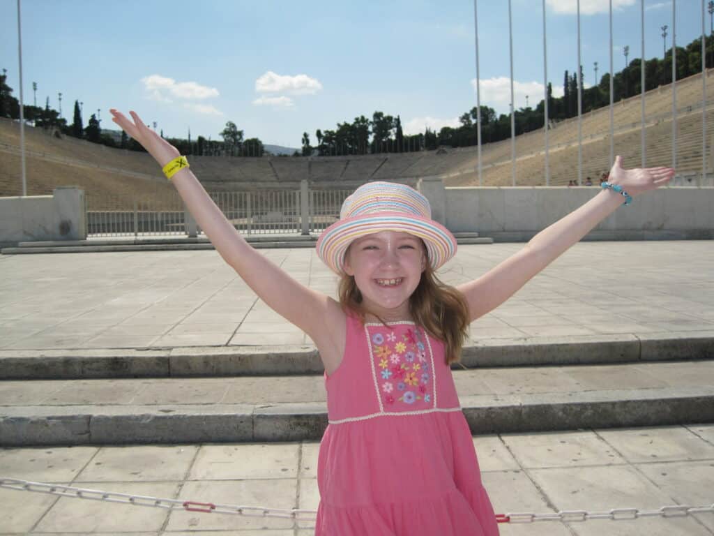 Young girl in pink dress and striped straw hat poses with outstretched arms in Olympic Stadium in Athens, Greece.