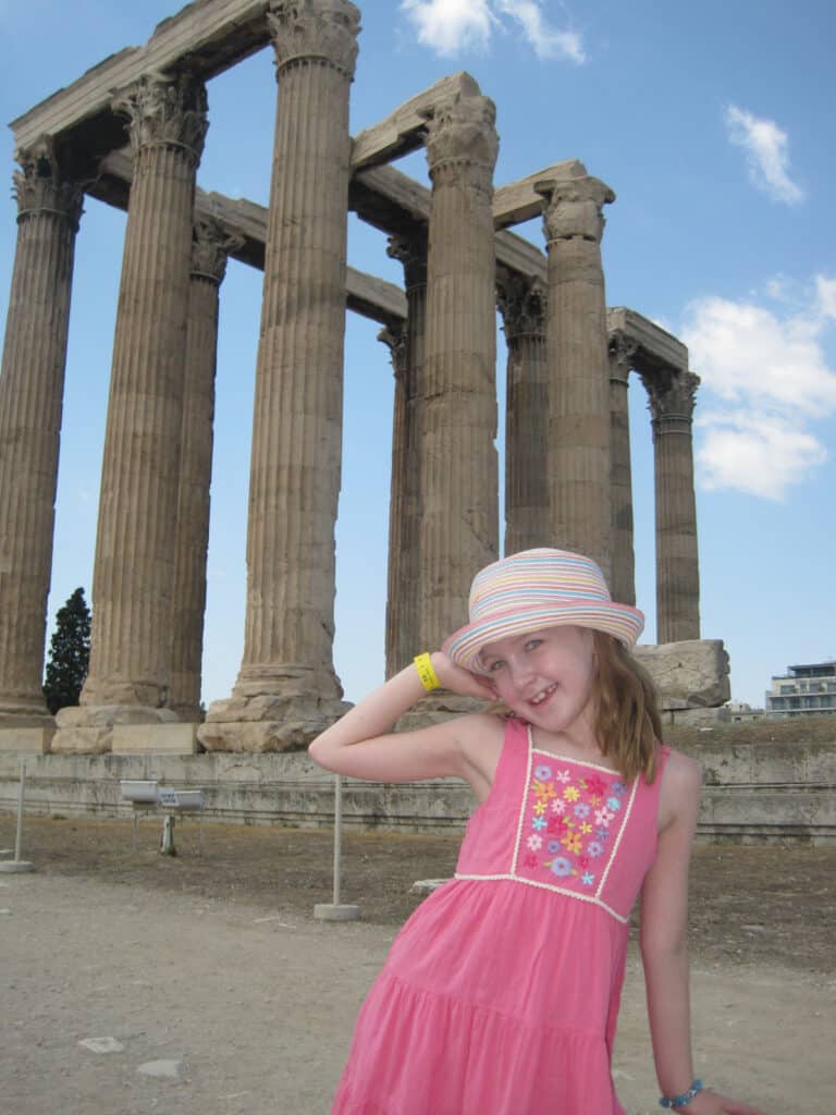 Smiling young girl in dark pink dress with embroidered flowers and striped hat poses with one hand on head in front of the remaining columns of the Temple of Olympian Zeus in Athens, Greece.