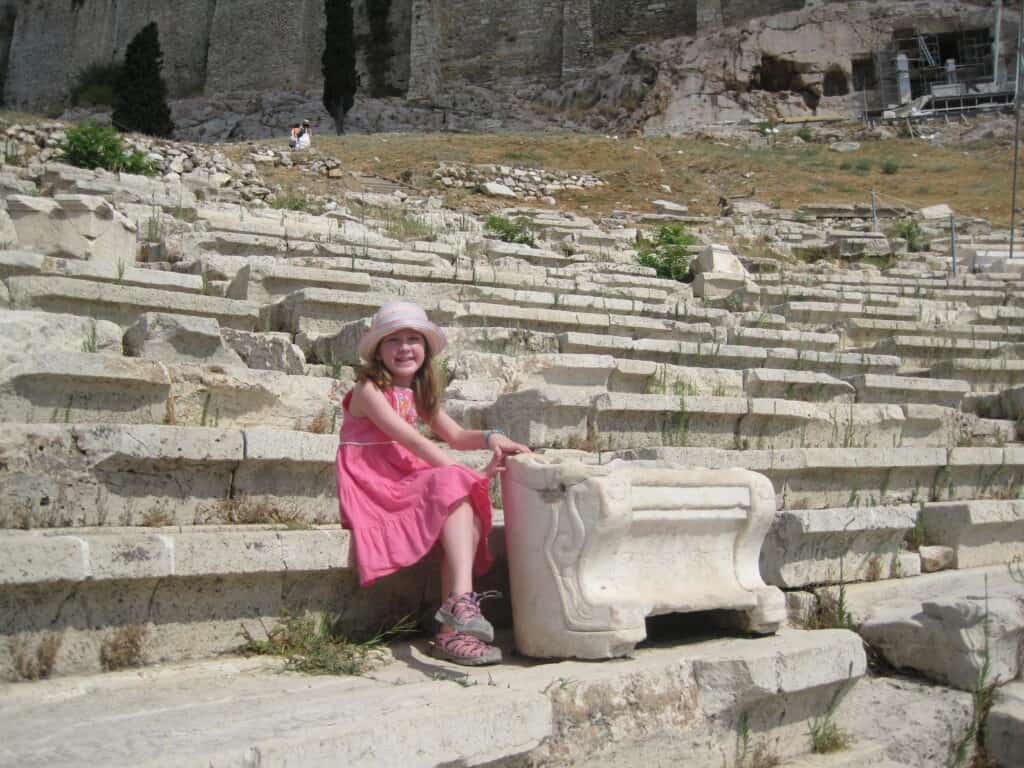 Young girl in pink dress and straw hat sitting on stone seats of the ruins of the Theatre of Dionysus.