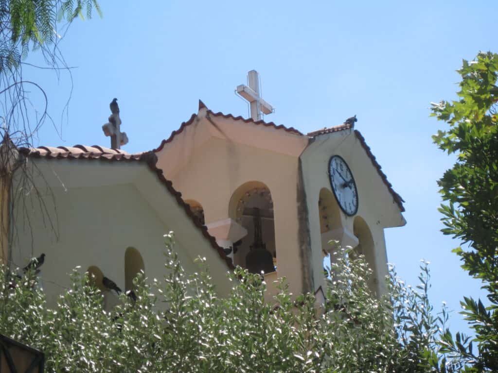 Top of church seen through trees with two crosses and a clock face.