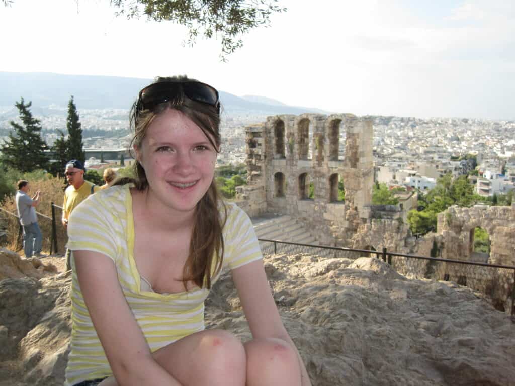 Teen girl in yellow and white t-shirt sitting on rocks with ruins in background on walk to Acropolis in Athens, Greece.