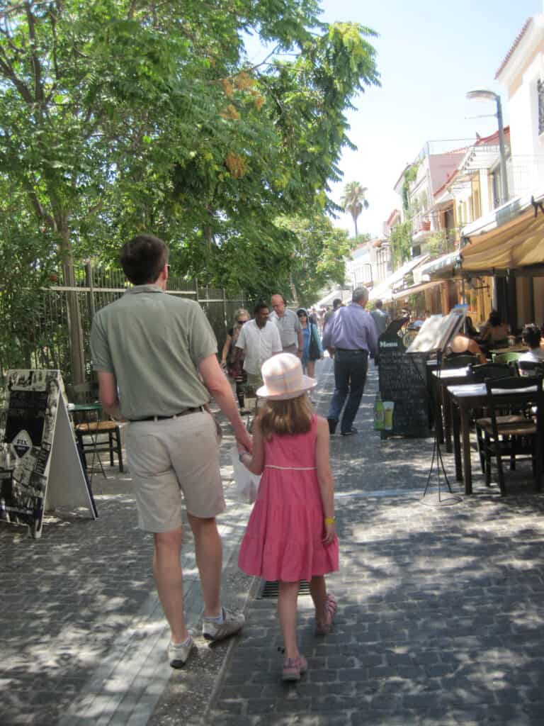 View from behind of dad and young girl walking through Plaka neighbourhood of Athens with restaurants and tables alongside.