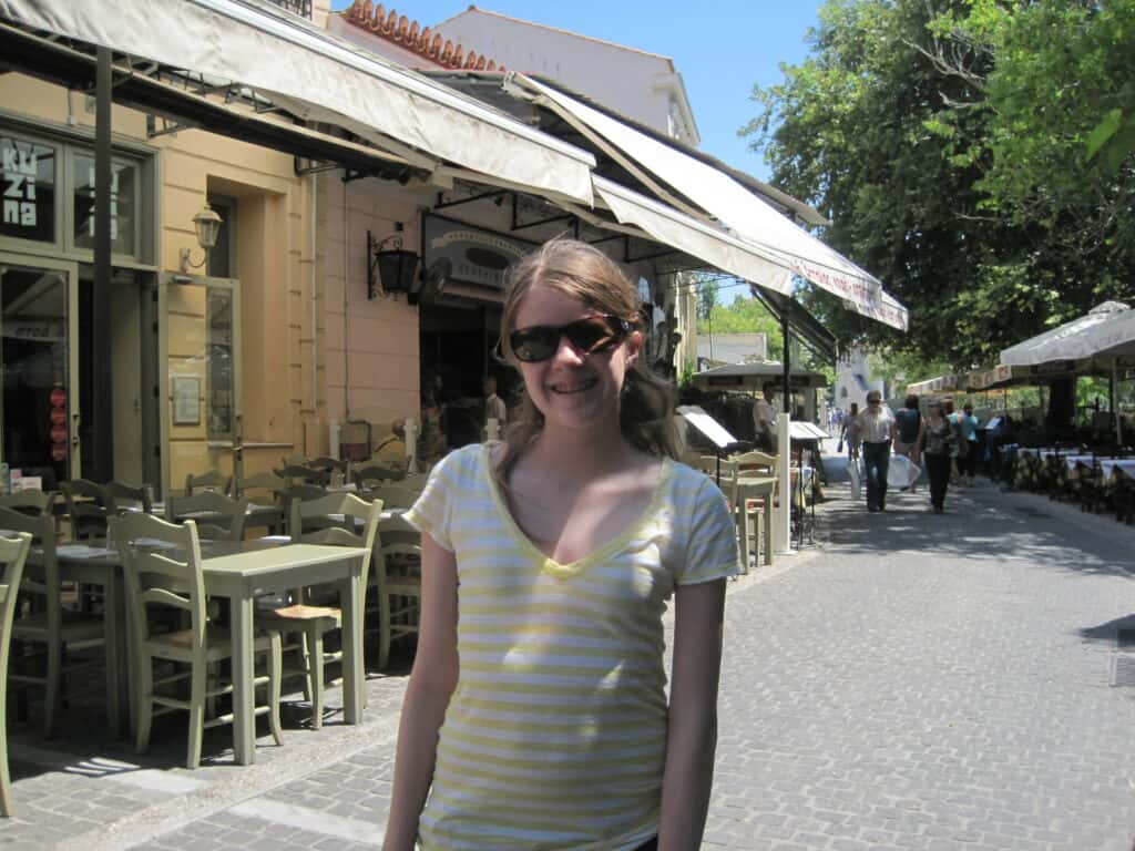 young teen girl in yellow and white striped shirt and sunglasses poses in front of restaurant in the plaka neighbourhood of Athens.