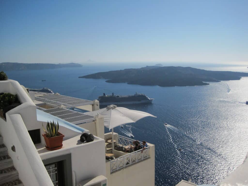 View past white building with people sitting on terrace under white umbrella to the Celebrity Equinox cruise ship anchored in the caldera off Santorini.