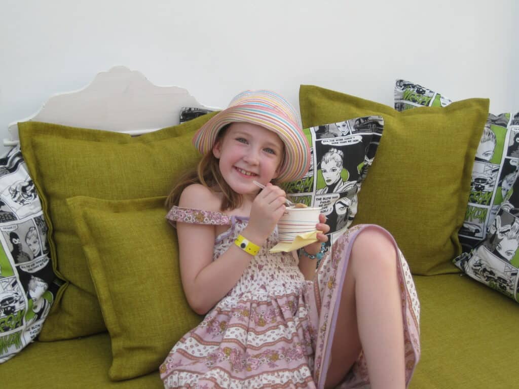 young girl in dress and straw hat sitting on sage green upholstered bench with matching sage green cushions and comic strip cushions eating cup of ice cream.