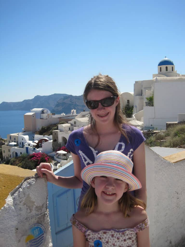 teen girl and younger girl standing in oia with white cliffside buildings behind them and cliffs and caldera beyond.