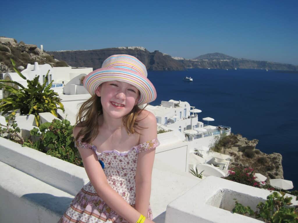 Young girl in flowered dress and striped multi-coloured straw hat sitting on white wall in Oia on Santorini with white buildings on cliff behind her and dark blue waters of the caldera.