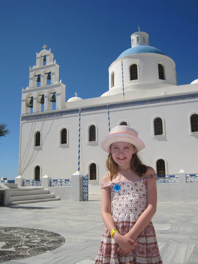 young girl in purple flowered sun dress and striped straw hat stands outside white church with blue dome and tier of bells in Oia on Santorini.