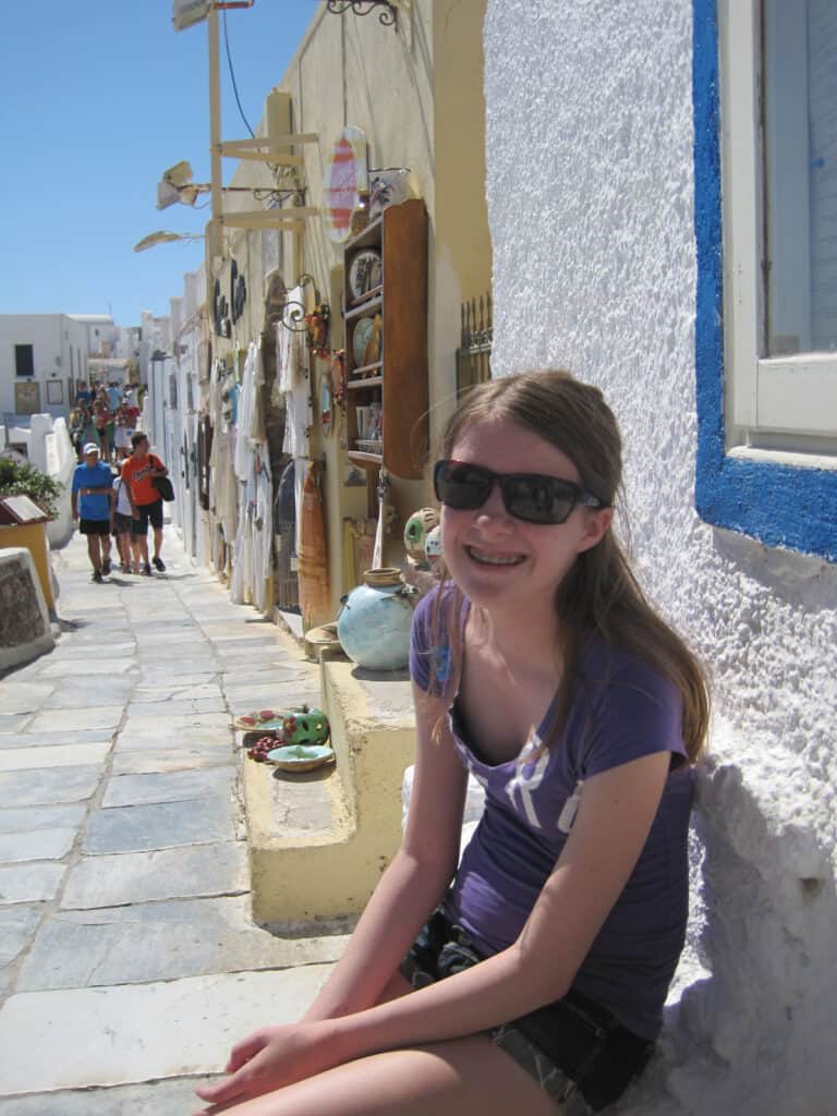 teen girl in purple t-shirt and sunglasses sitting outside shop in oia with street of shops displaying merchandise and shoppers walking in background.