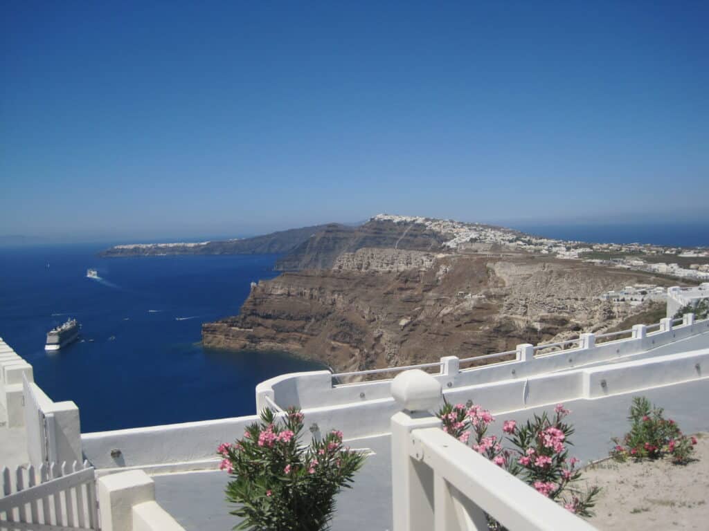 white balcony of building with plants with pink flowers and view of caldera with cruise ships and cliffs of island.