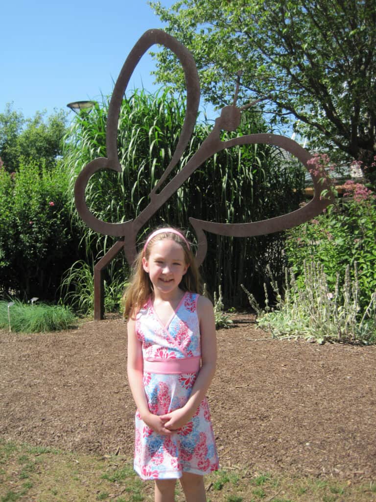 Young girl in pink and blue patterned dress standing by wrought iron sculpture at entrance to Niagara Parks Butterfly Conservatory.