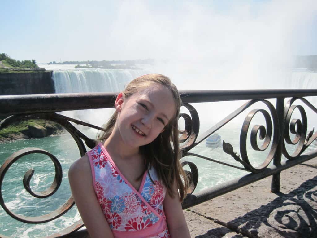 young girl in pink patterned dress standing by black railing with niagara falls and boat in background.