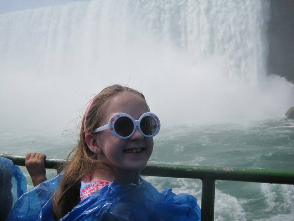 young girl in blue poncho and round white sunglasses turns to look at camera with churning waters and horseshoe falls in background, Niagara Falls, Canada.