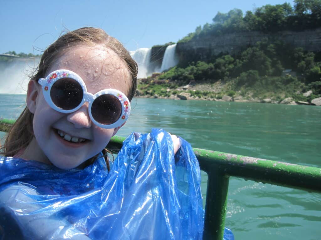 Smiling young girl wearing blue poncho and round white sunglasses with water dripping down face on Maid of the Mist with American Falls in background.