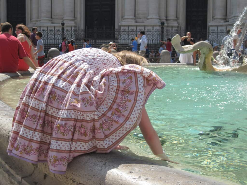 young girl in light purple flowered dress squats on edge of fountain playing in the water in rome.