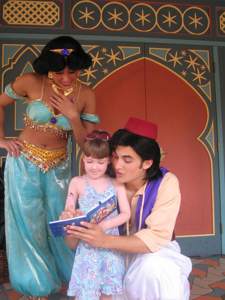 Aladdin kneeling with young girl and signing her autograph while Jasmine looks on at Magic Kingdom, Disney World.