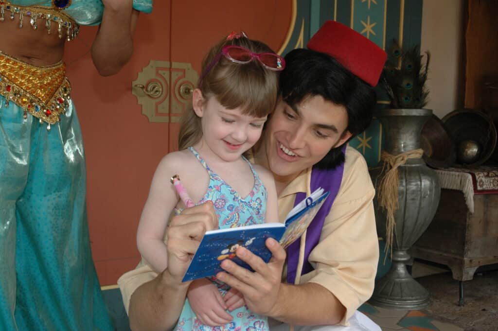 Young girl and Aladdin smiling as he signs her autograph book at Disney World.