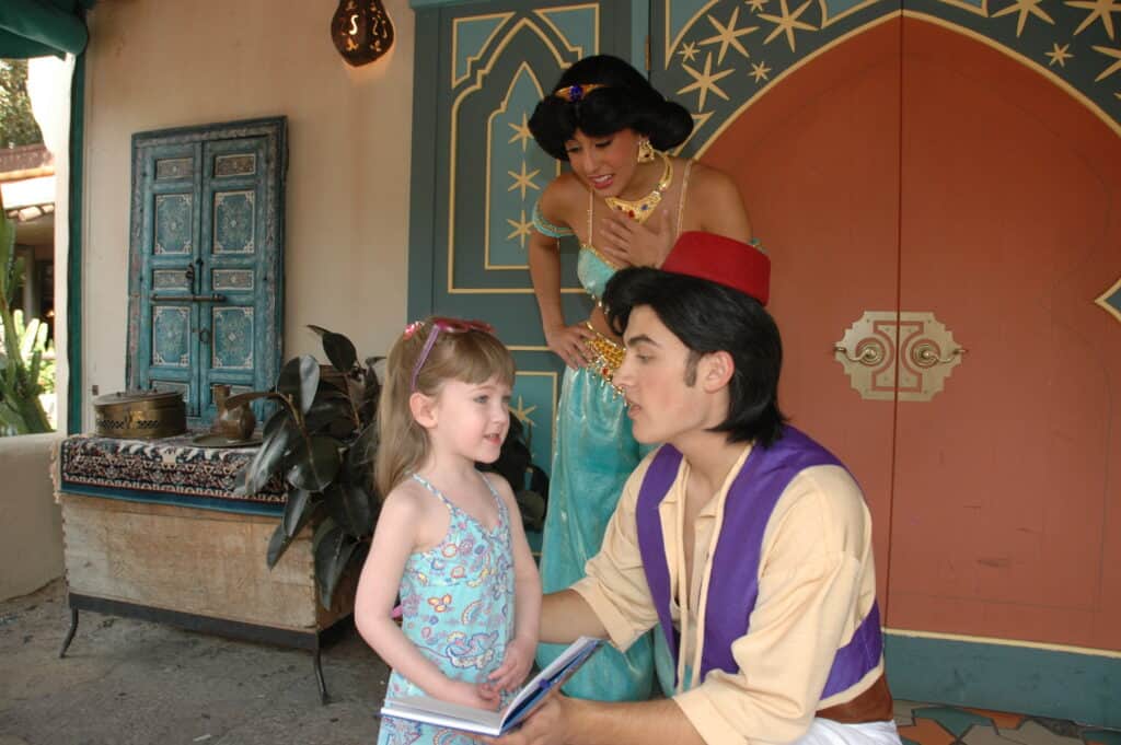 A kneeling Aladdin talks to young girl while Jasmine listens.