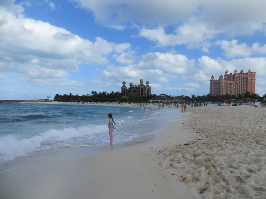 Young girl standing at edge of ocean on Paradise Beach at Atlantis Resort in the Bahamas with resort towers in background.