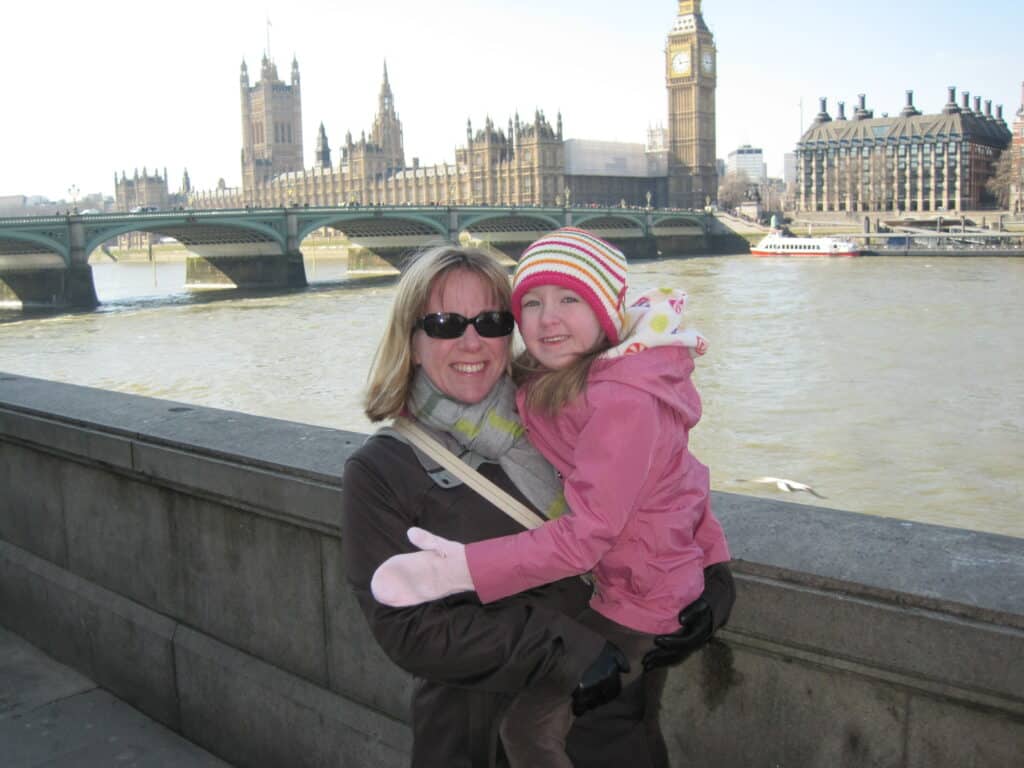 Woman wearing sunglasses holding young girl wearing pink coat, mittens and striped hat beside the Thames with bridge and Big Ben in background.