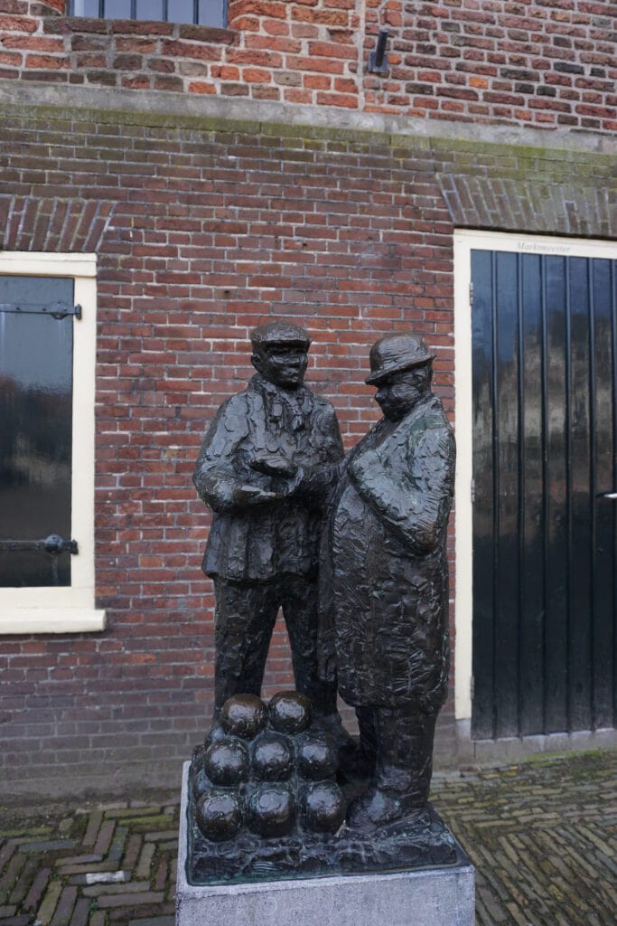 Alkmaar Cheese Market bronze sculpture in front of red brick wall with two men wearing hats haggling over price of cheese stacked at their feet.