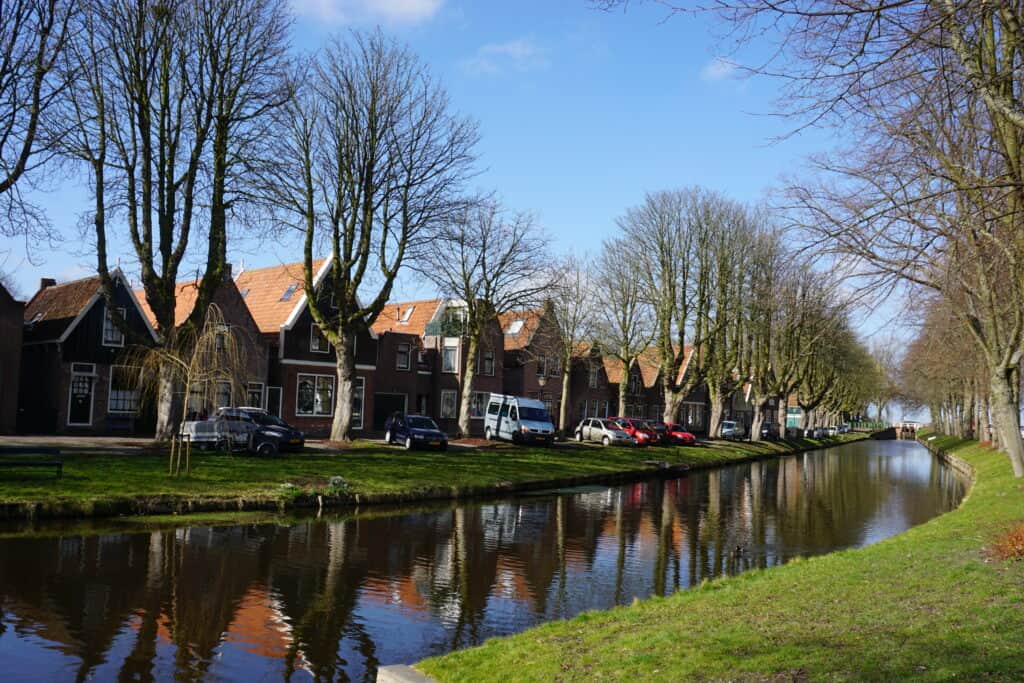 A row of homes with cars parked in front alongside tree-lined canal in Edam with reflections in the water.