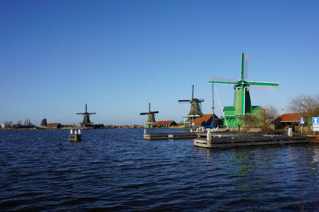 Windmill park at Zaanse Schans - four windmills alongside water - green windmill in foreground and three grey/brown coloured windmills in background.