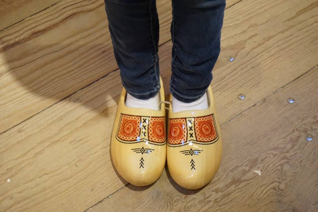 Child's legs in dark jeans and white socks wearing light-coloured wooden clogs with red and black pattern.