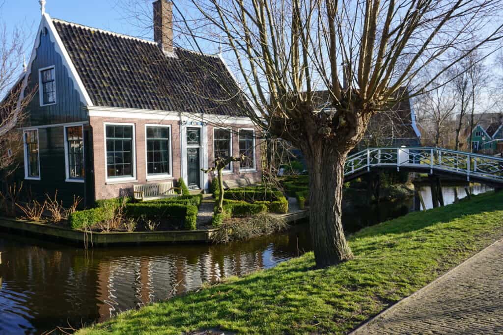 Bridge with railing over narrow canal to small house with many windows and garden with hedges at Zaanse Schans.