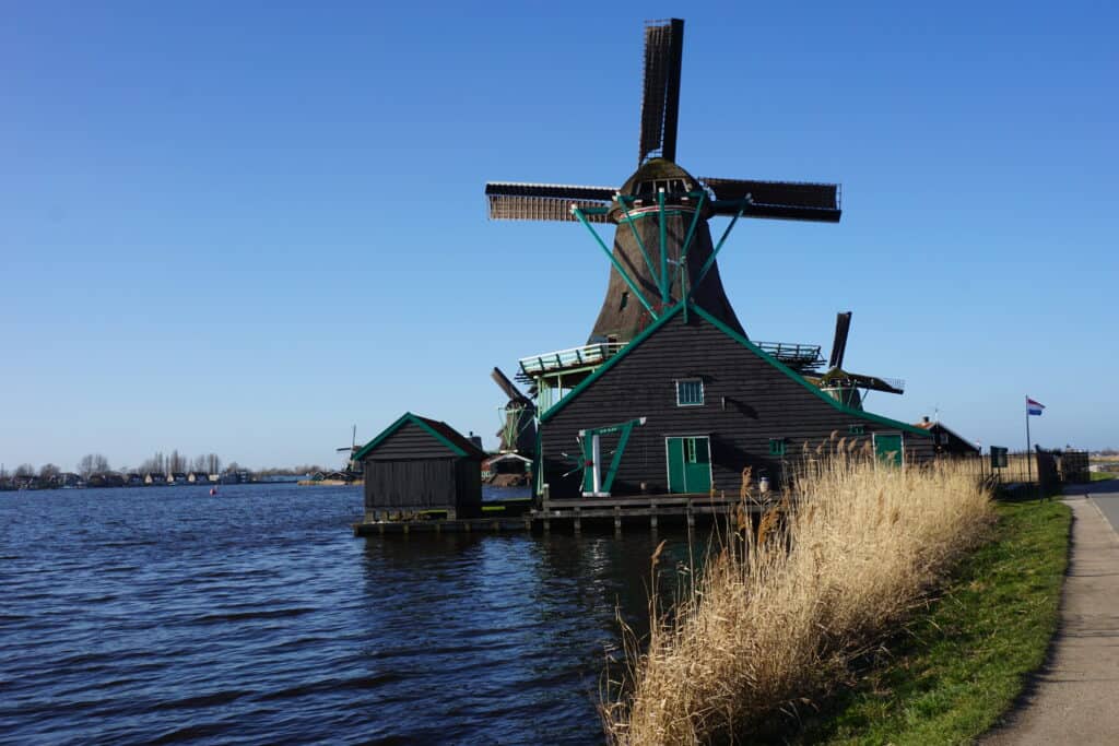 Weathered grey/brown windmill and buildings with green trim and doors sitting on wooden platform over water alongside path and grasses at Zaanse Schans on spring day with bright blue sky.