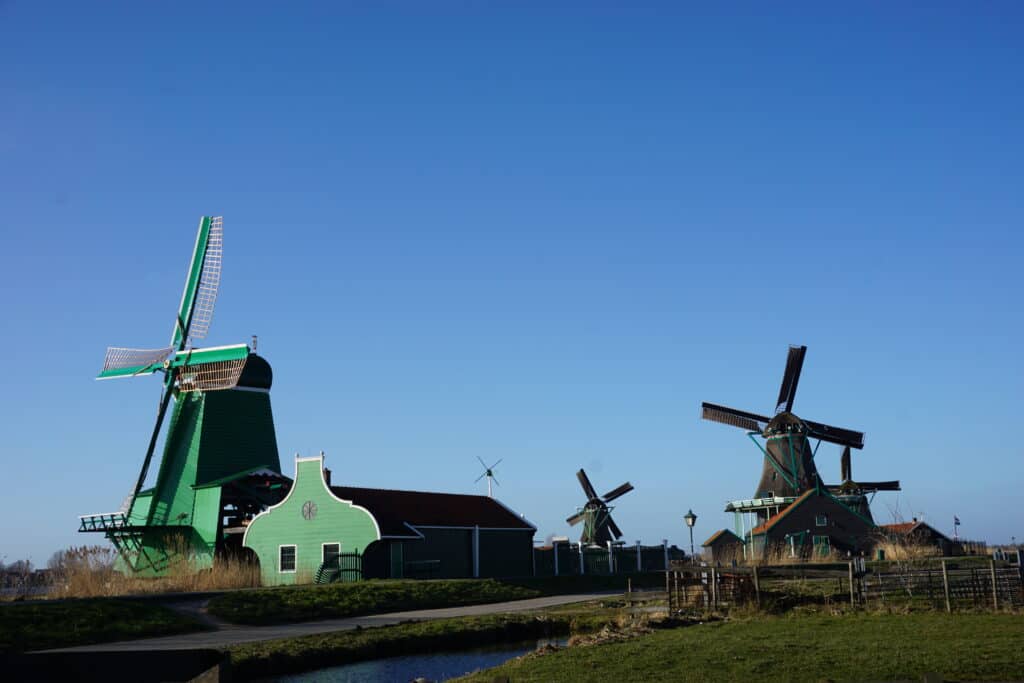 Zaanse Schans Windmill Park with bright blue sky - green windmill and building in foreground alongside canal with two weathered grey windmills in background.