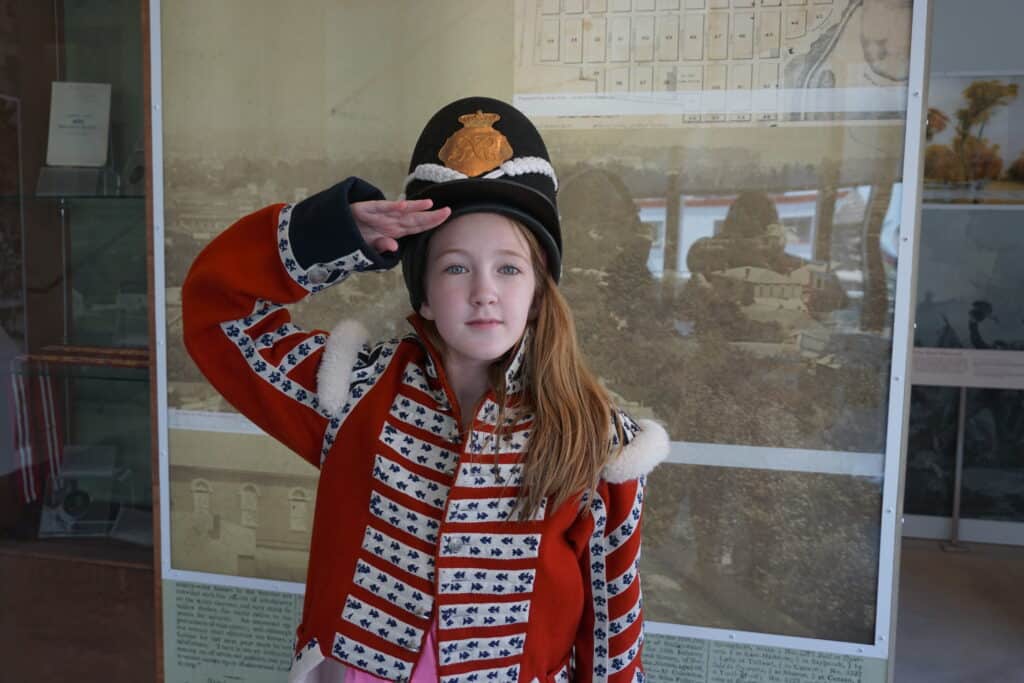 Young girl salutes while wearing red and white military jacket and black hat at Niagara Falls History Museum.