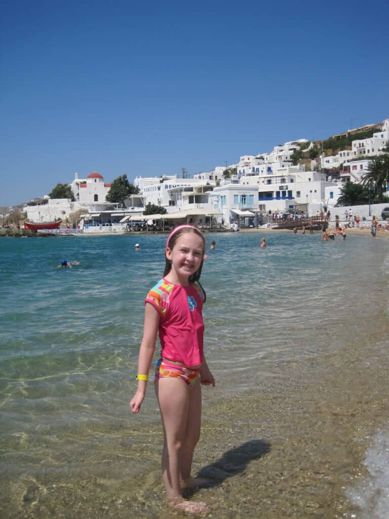 Young girl in bathing suit standing in water on Mykonos with white and blue buildings in background.